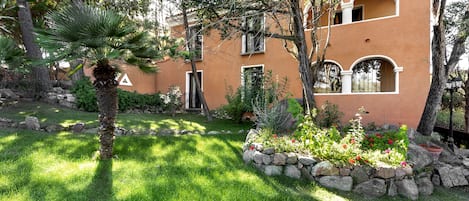 Un edificio de dos pisos con fachada de estuco, cimientos de piedra y un jardín cuidado con una palmera y plantas con flores.