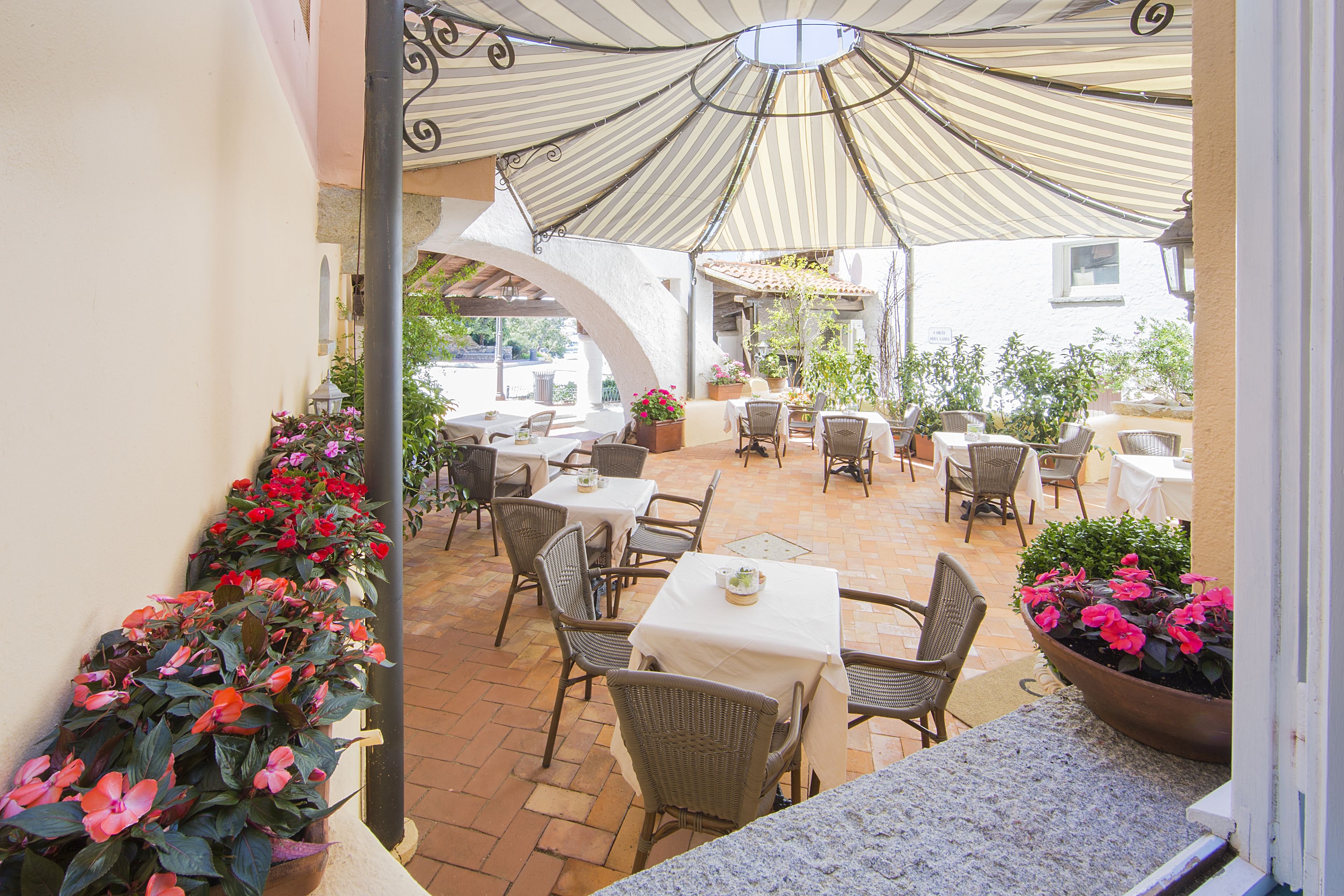 An outdoor patio area with tables and chairs, surrounded by potted plants and a striped awning.