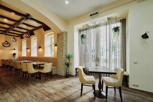 Un restaurante con suelo de madera, pared de piedra y un amplio ventanal con cortinas. Se observan mesas y sillas dispuestas para comer.