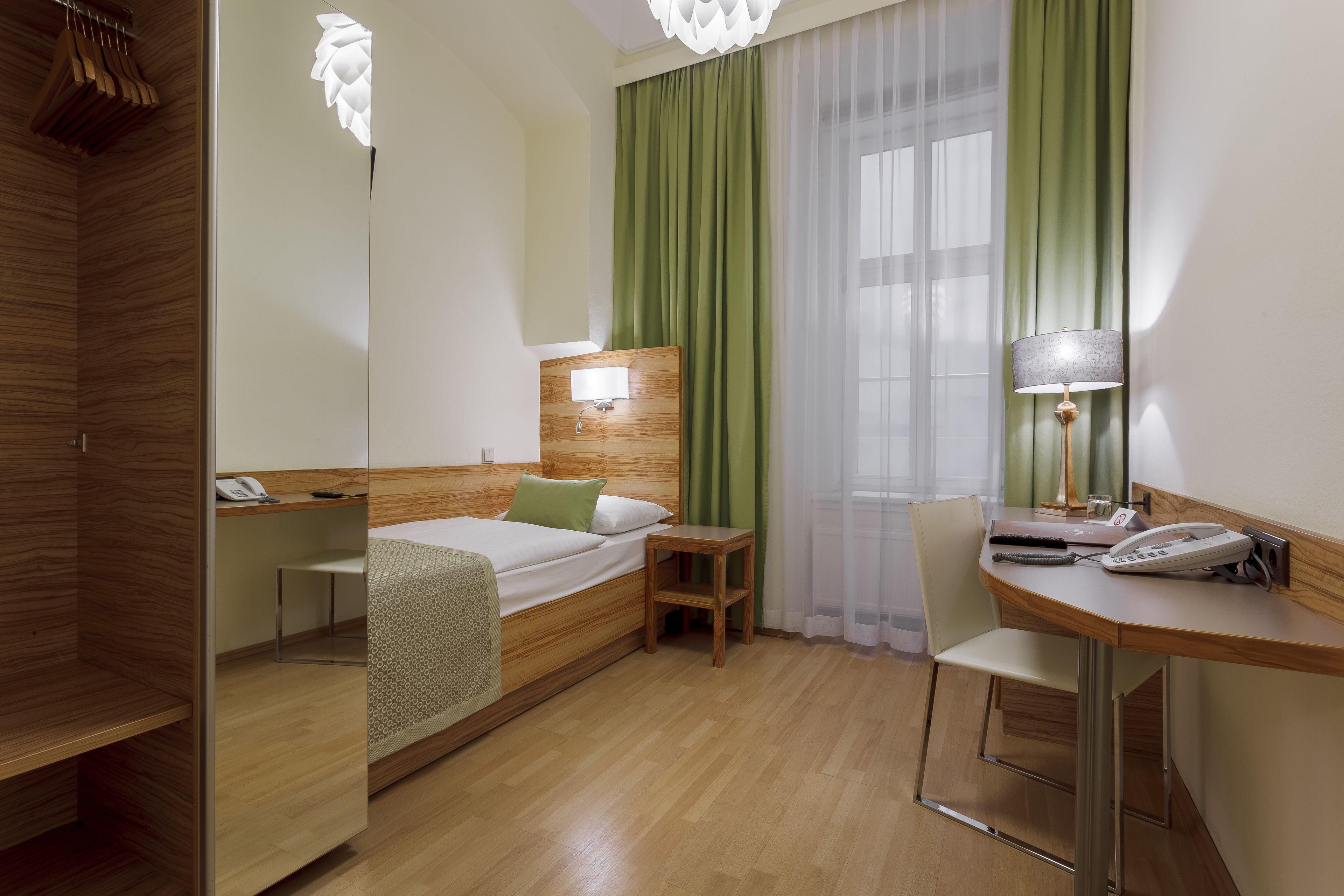 A hotel room with a bed, bedside table, lamp, chair, and a desk with a phone.