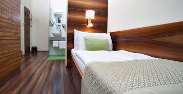 A hotel room with a wooden floor, a bed with a patterned bedspread, a bedside lamp, and a bathroom with a sink and mirror.
