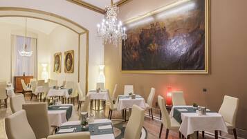 A dining area with tables set for four, a chandelier, and a large painting on the wall.