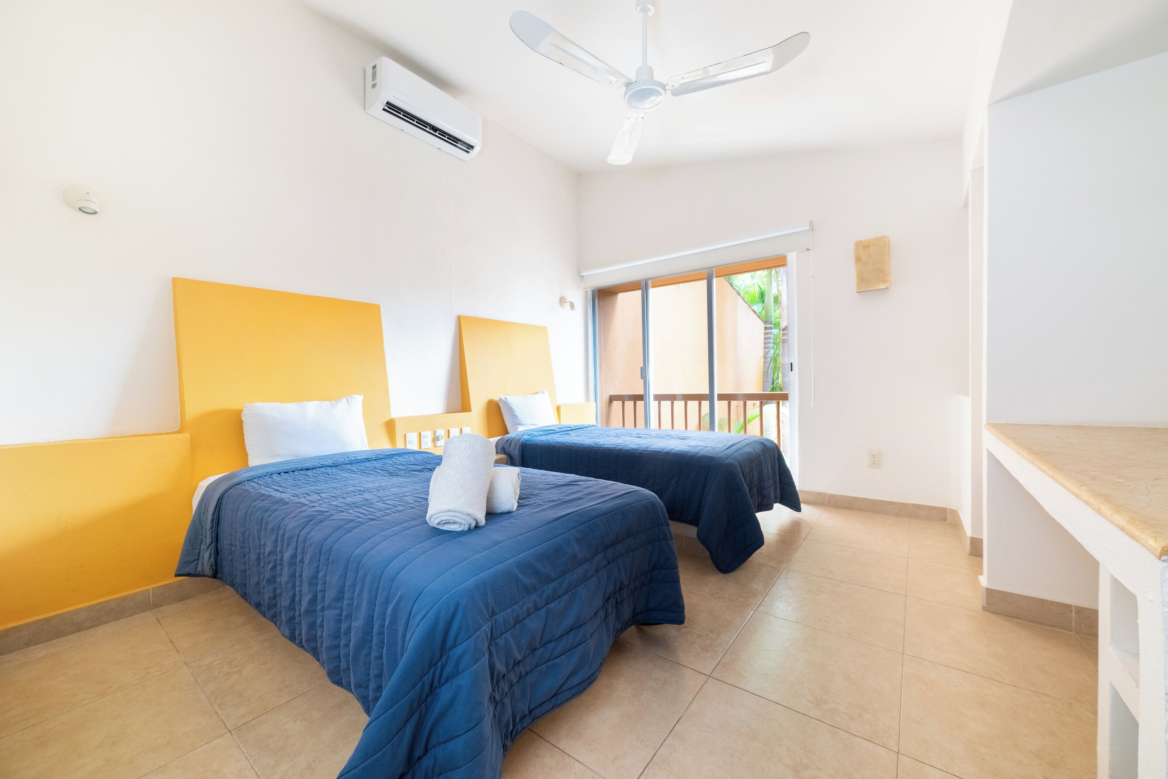 Habitación de hotel con dos camas, ventilador de techo, aire acondicionado y ventana con vistas a la vegetación.