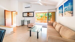 A modern living room with a glass coffee table, a sofa, a ceiling fan, and a view of palm trees through a glass door. - Hotel Ixzi Plus (Ixtapa)