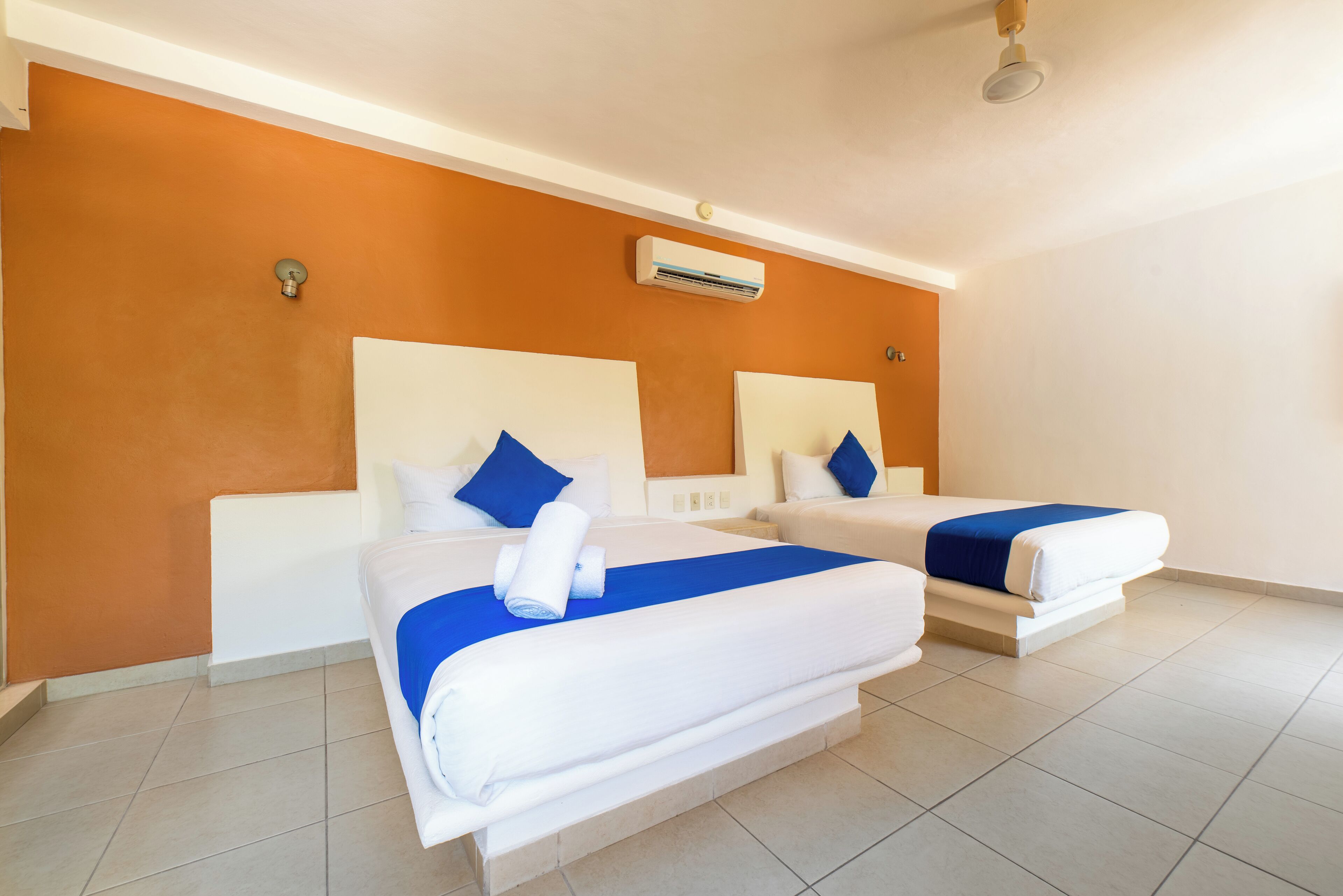 A hotel room with two beds, white bedding, blue accents, an air conditioning unit, and a tiled floor.