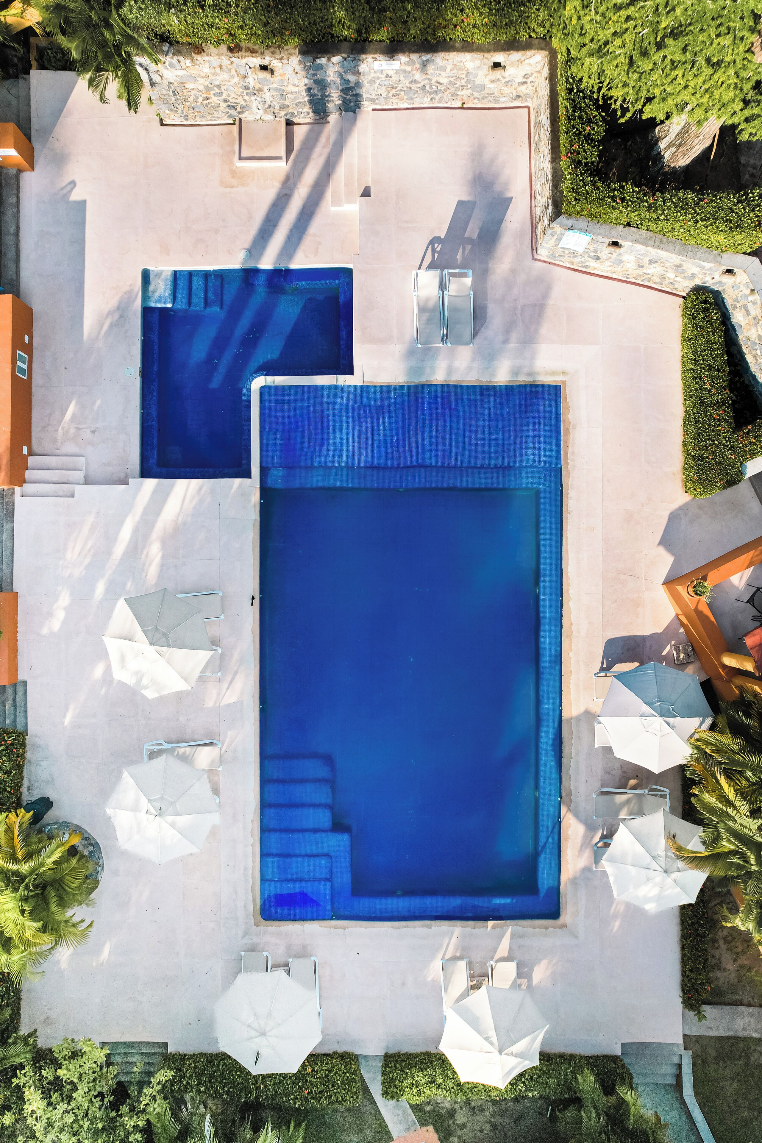 An aerial view of a swimming pool area with two pools, surrounding white umbrellas, and a stone wall.