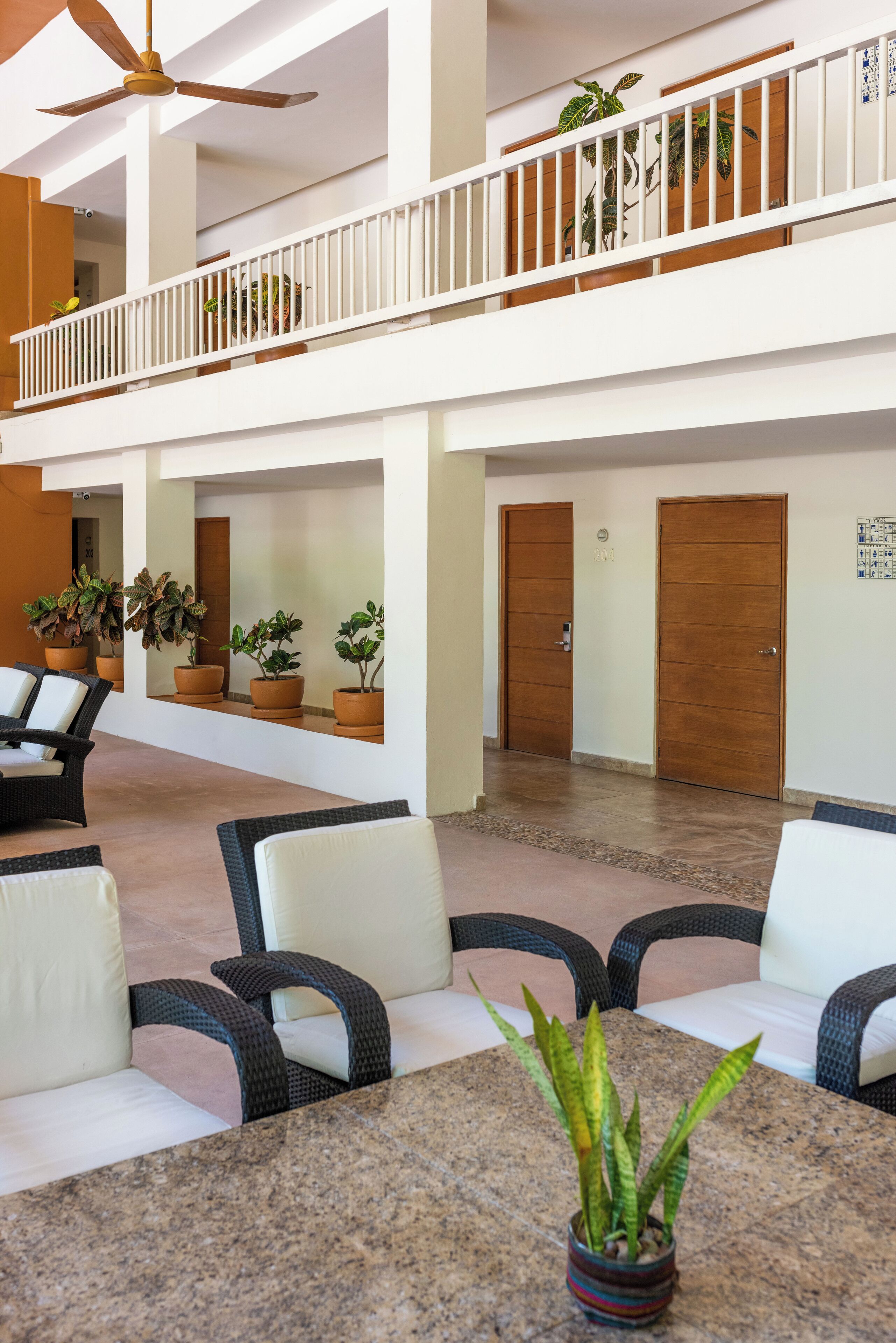 Un hall d’entrée spacieux avec un plafond haut, un ventilateur de plafond et un balcon doté de balustrades blanches. On y trouve plusieurs plantes en pot ainsi qu’un coin salon avec des fauteuils en osier et une table.