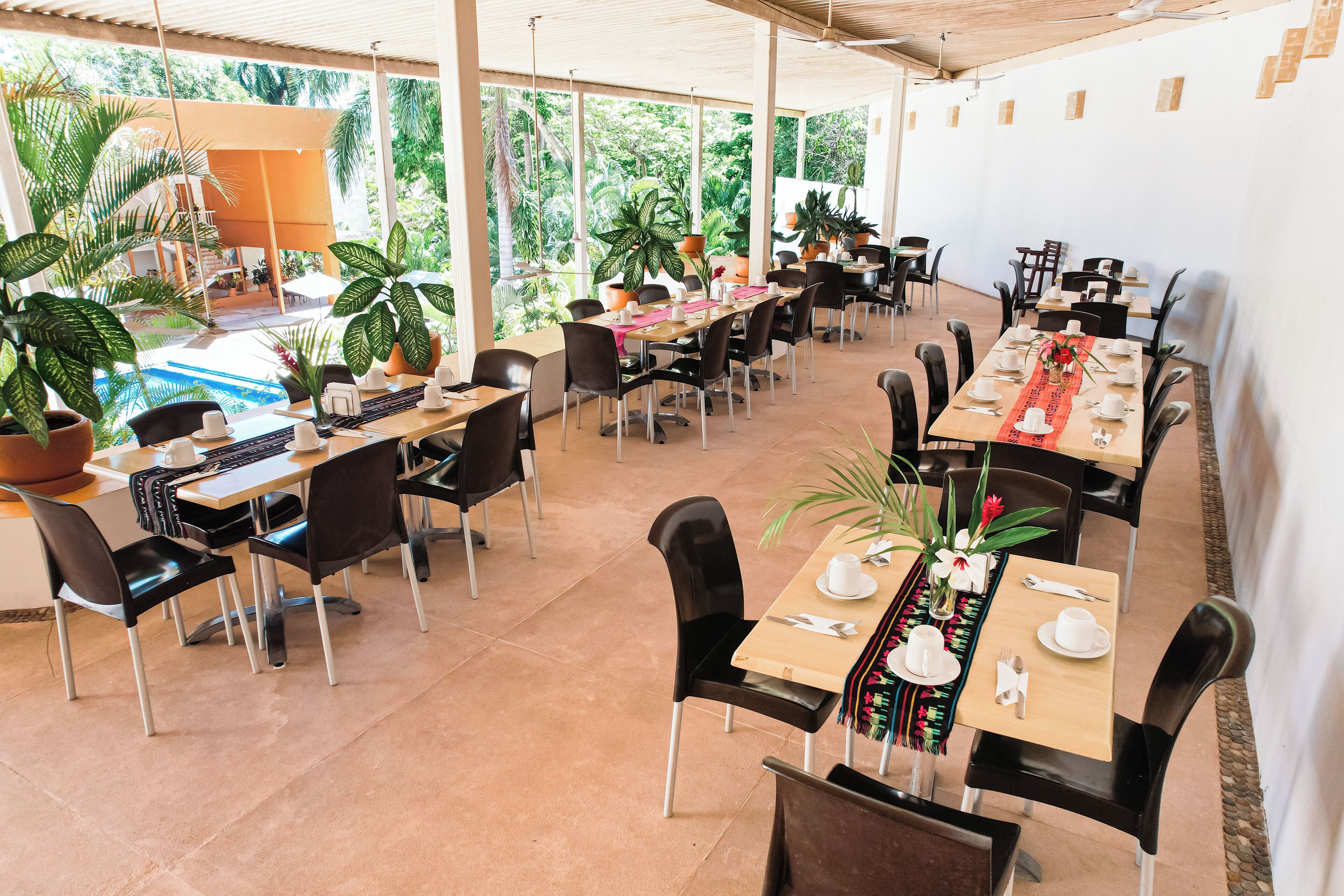 A spacious outdoor dining area with multiple tables and chairs, surrounded by potted plants and large windows offering views of the lush greenery outside.