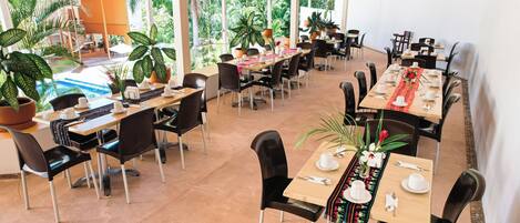 A spacious outdoor dining area with multiple tables and chairs, surrounded by potted plants and large windows offering views of the lush greenery outside.