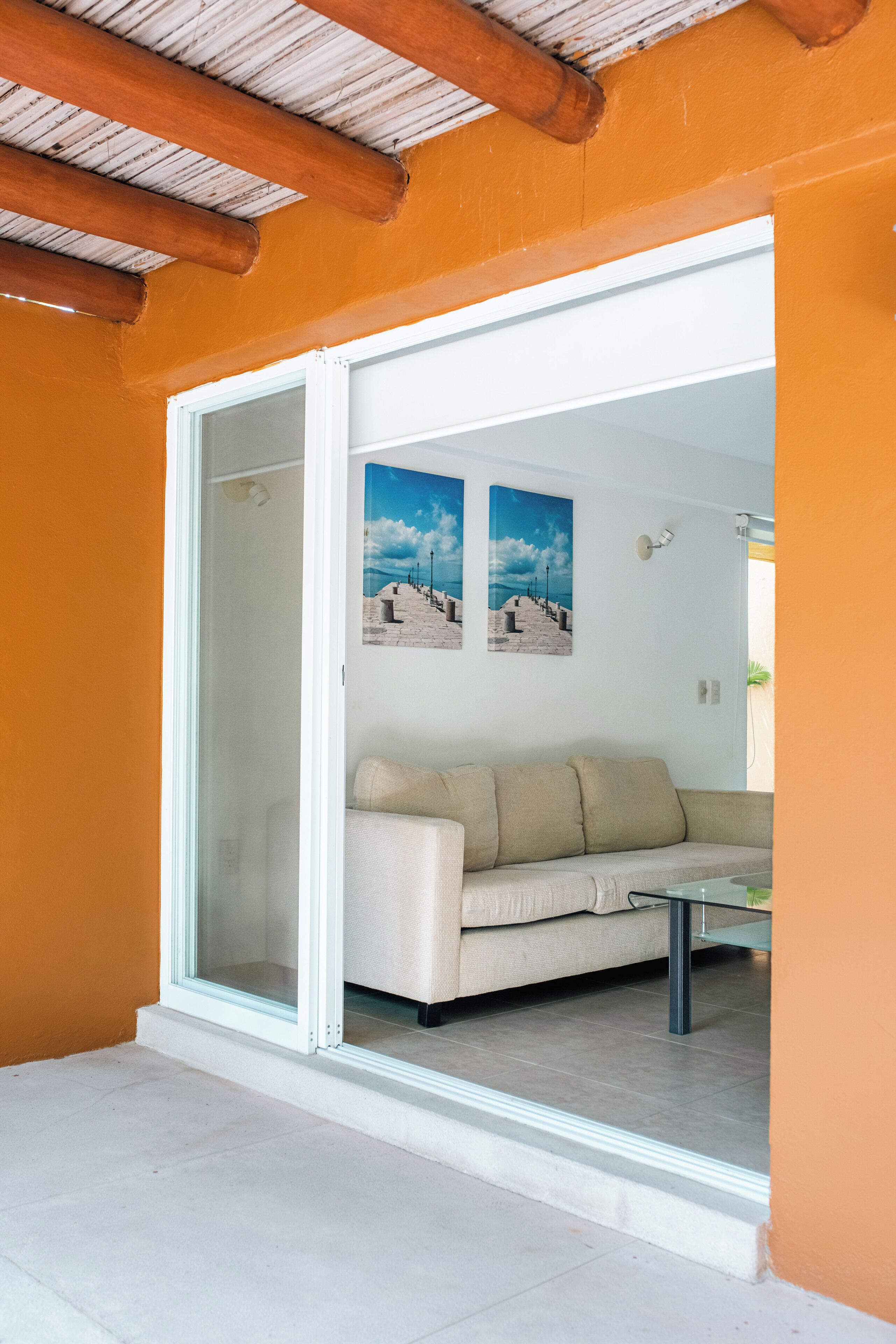 Una habitación con puerta de cristal corredera que da a un balcón, un sofá color beige y dos cuadros en la pared.