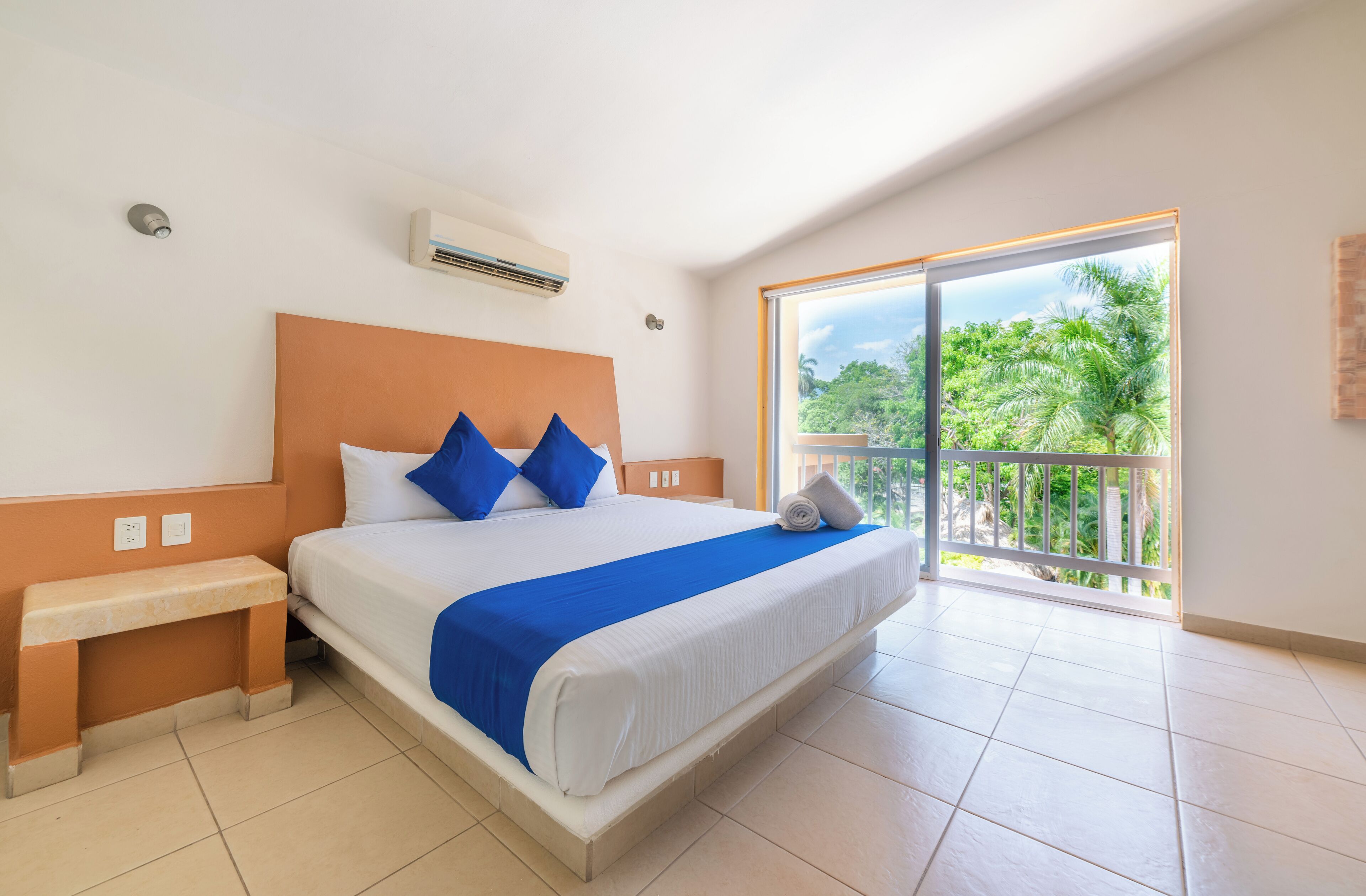 Une chambre d’hôtel avec un grand lit, un banc, une fenêtre donnant sur un espace verdoyant et un appareil de climatisation.