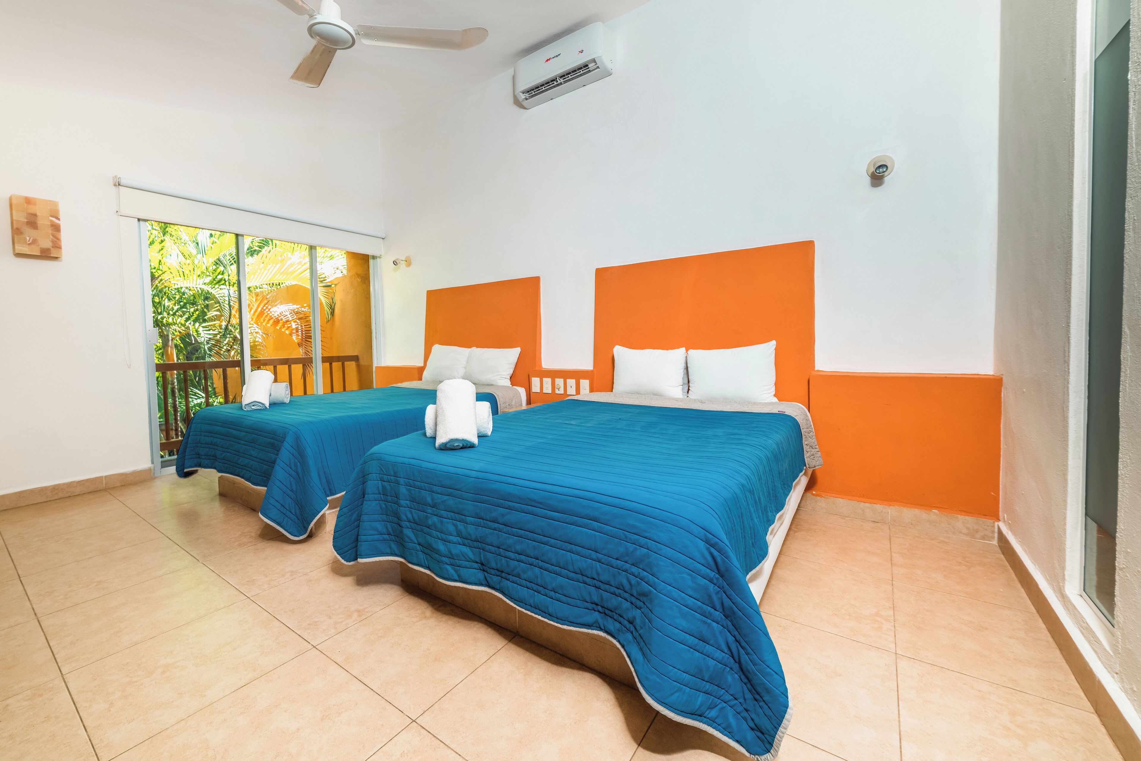 A hotel room with two beds, each with an orange headboard and blue bedding. There is a ceiling fan and an air conditioning unit visible.