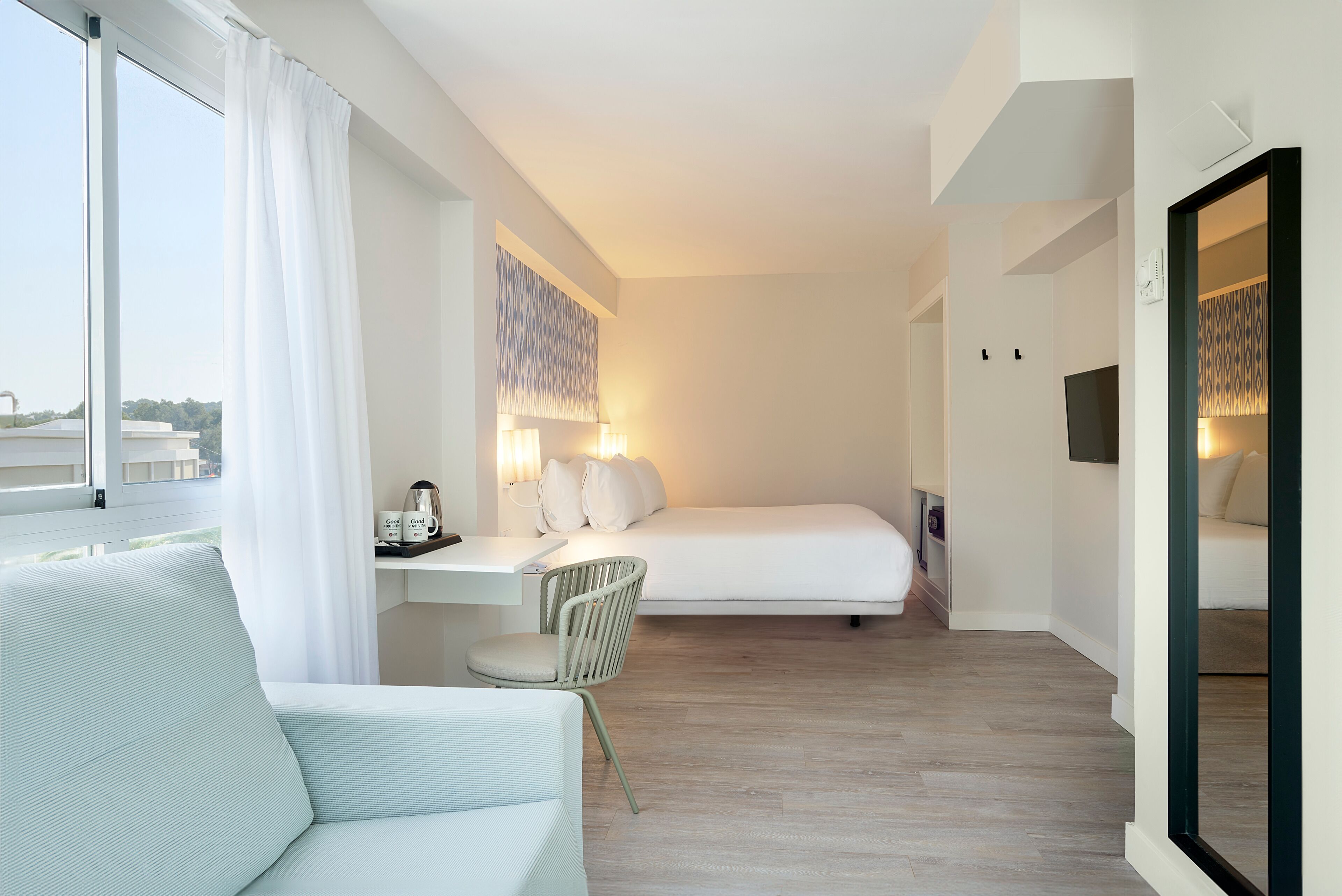 Una habitación de hotel moderna con un ventanal grande, una cama, un escritorio con una silla y una mesita de noche con una lámpara.