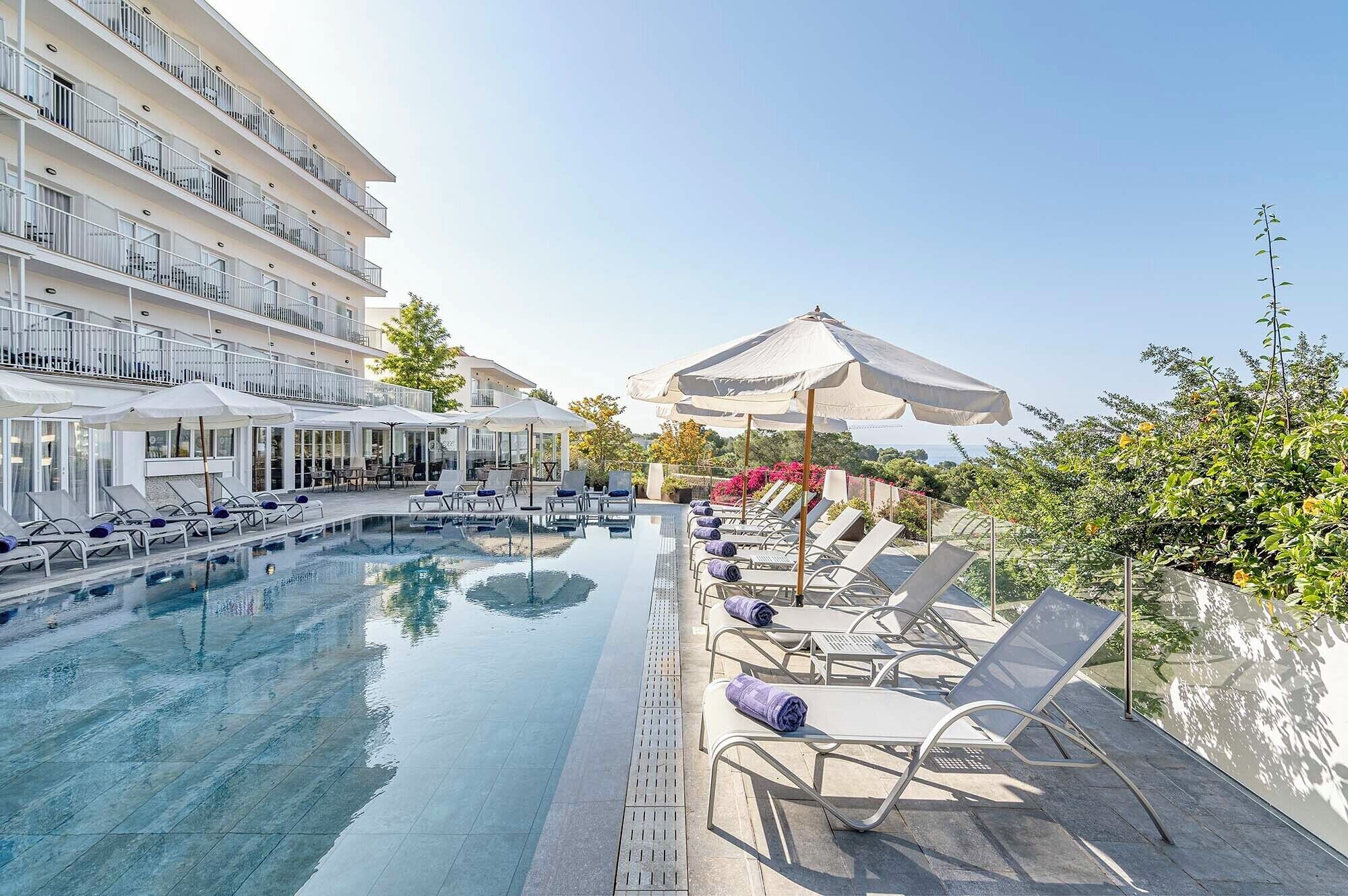 Una piscina con sillas de descanso y sombrillas, rodeada por un edificio de varios pisos con balcones.