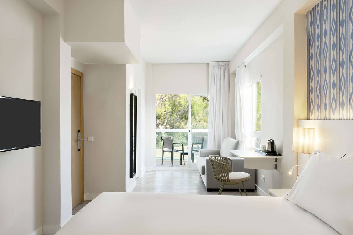 A modern hotel room with a large bed, a desk with a chair, a TV, and a view of the outdoors.