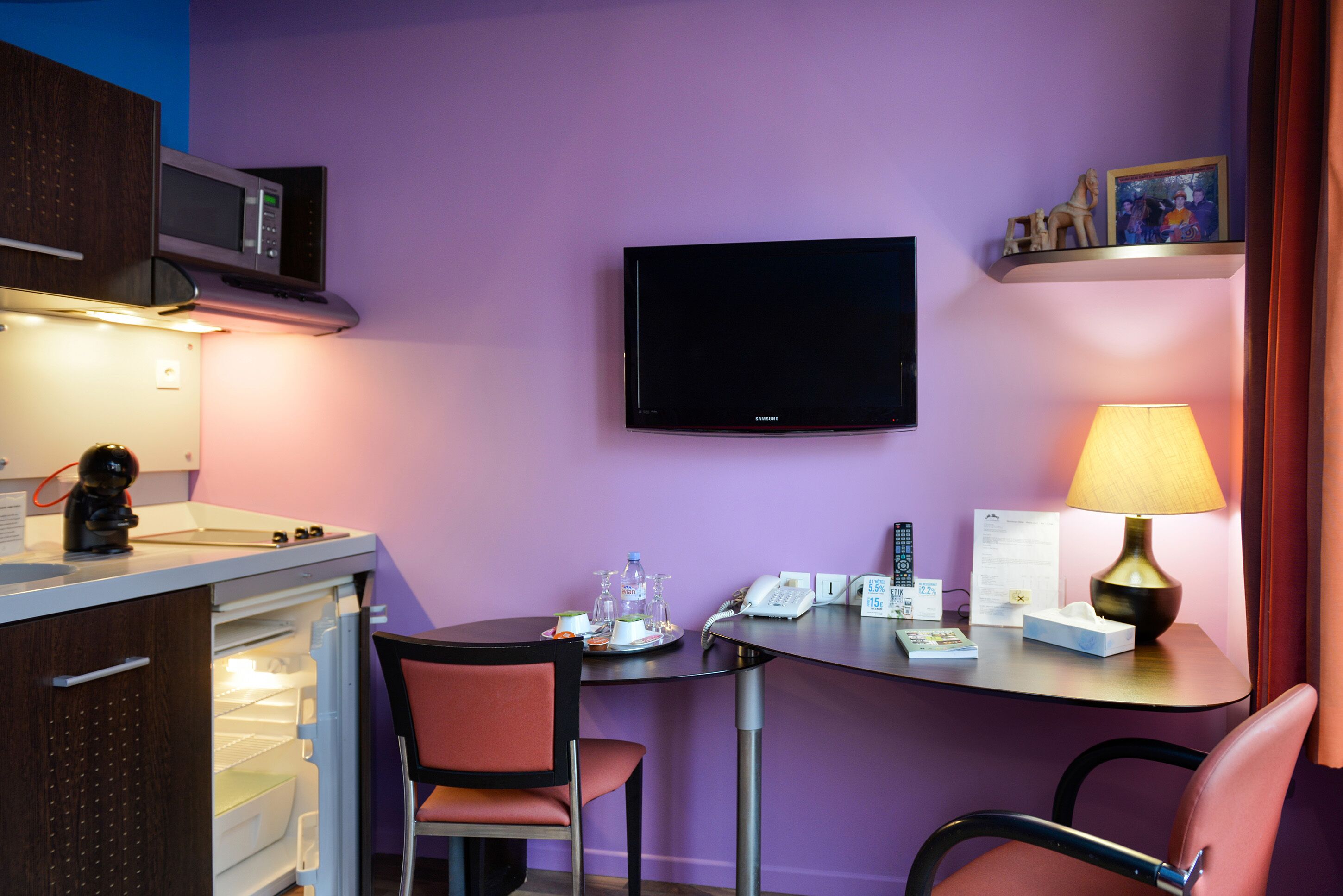 Habitación de hotel compacta con kitchenette, un televisor de pantalla plana, una mesita con lámpara y dos sillas.