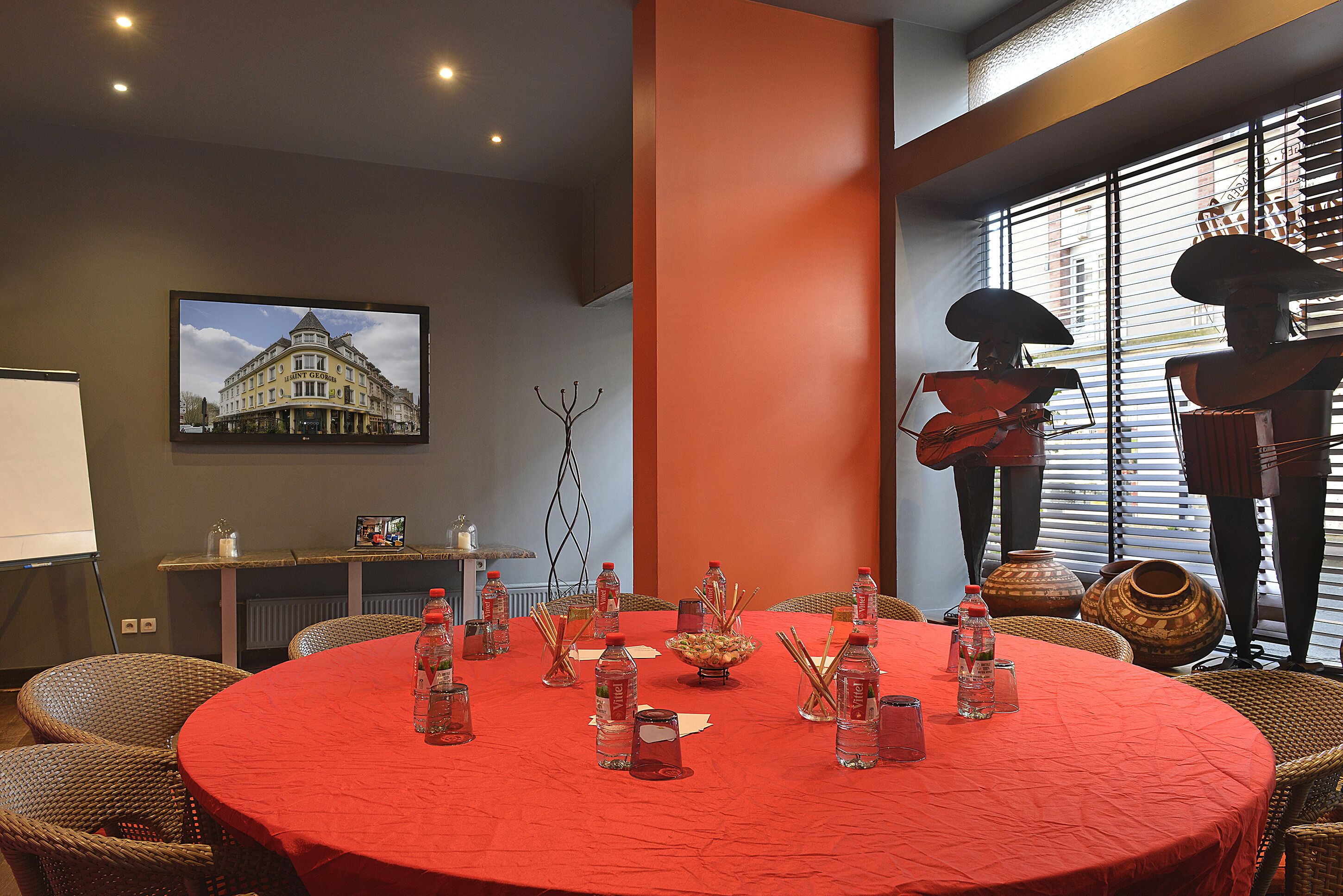 A conference room with a red table, wicker chairs, a projector screen, and a large window with blinds.