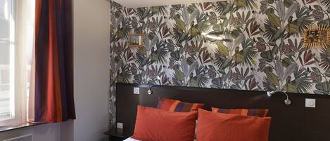 Un dormitorio con cama, ventana con cortinas, pared con estampado de hojas tropicales y mesita de noche con lámpara.