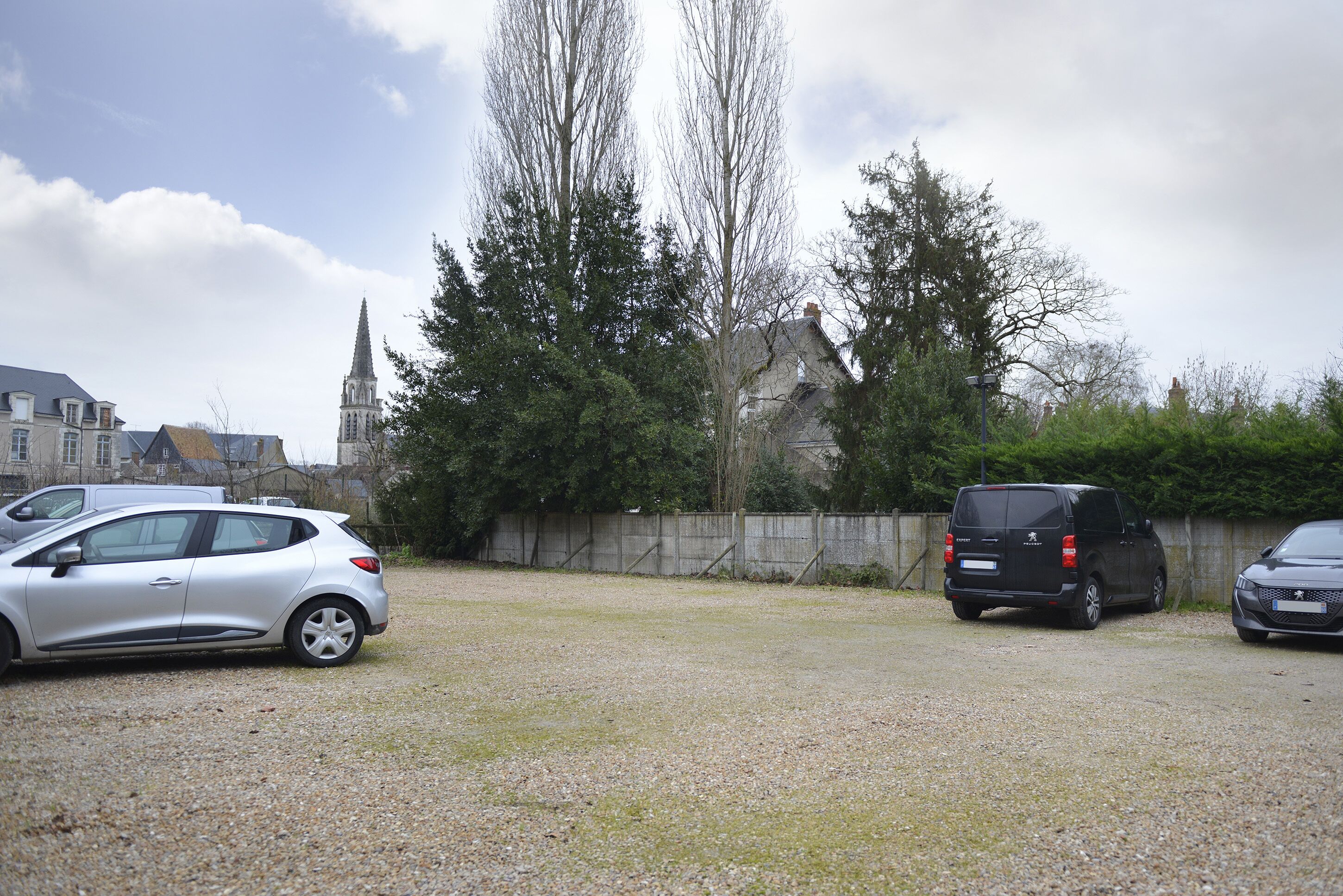 A parking area with two cars, a gravel surface, and a church with a tall spire in the background.