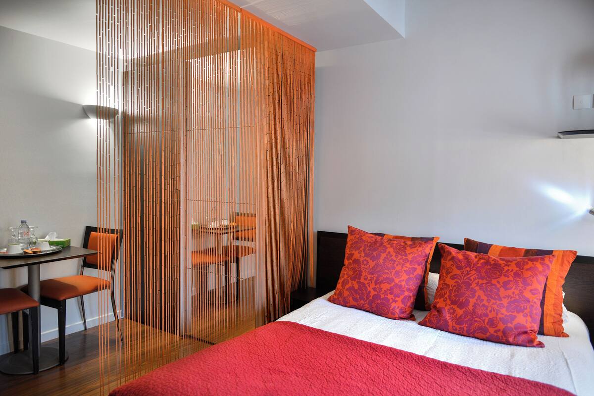 A hotel room with a bed, orange pillows, a red blanket, a dining area with a table and chairs, and a partition with vertical lines.
