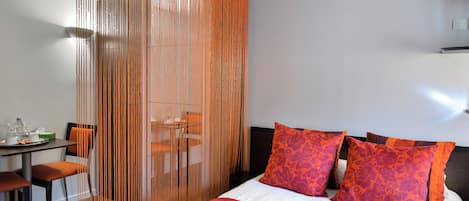 Habitación de hotel con cama, almohadones naranja, una colcha roja, zona de comedor con mesa y sillas, y un separador con líneas verticales.