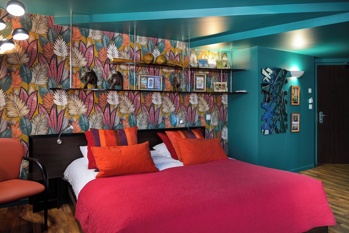A bedroom with a bed, a chair, a wall with a leaf pattern, and shelves with various items.