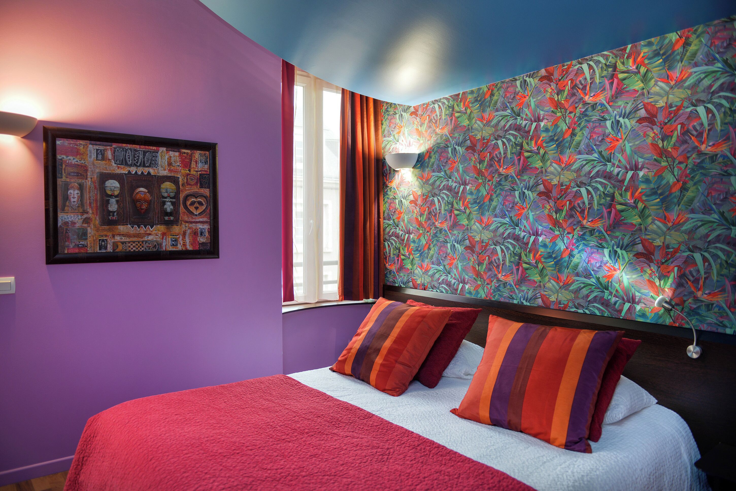 A bedroom with a purple wall, a floral wallpaper, a framed artwork, and a bed with striped pillows.