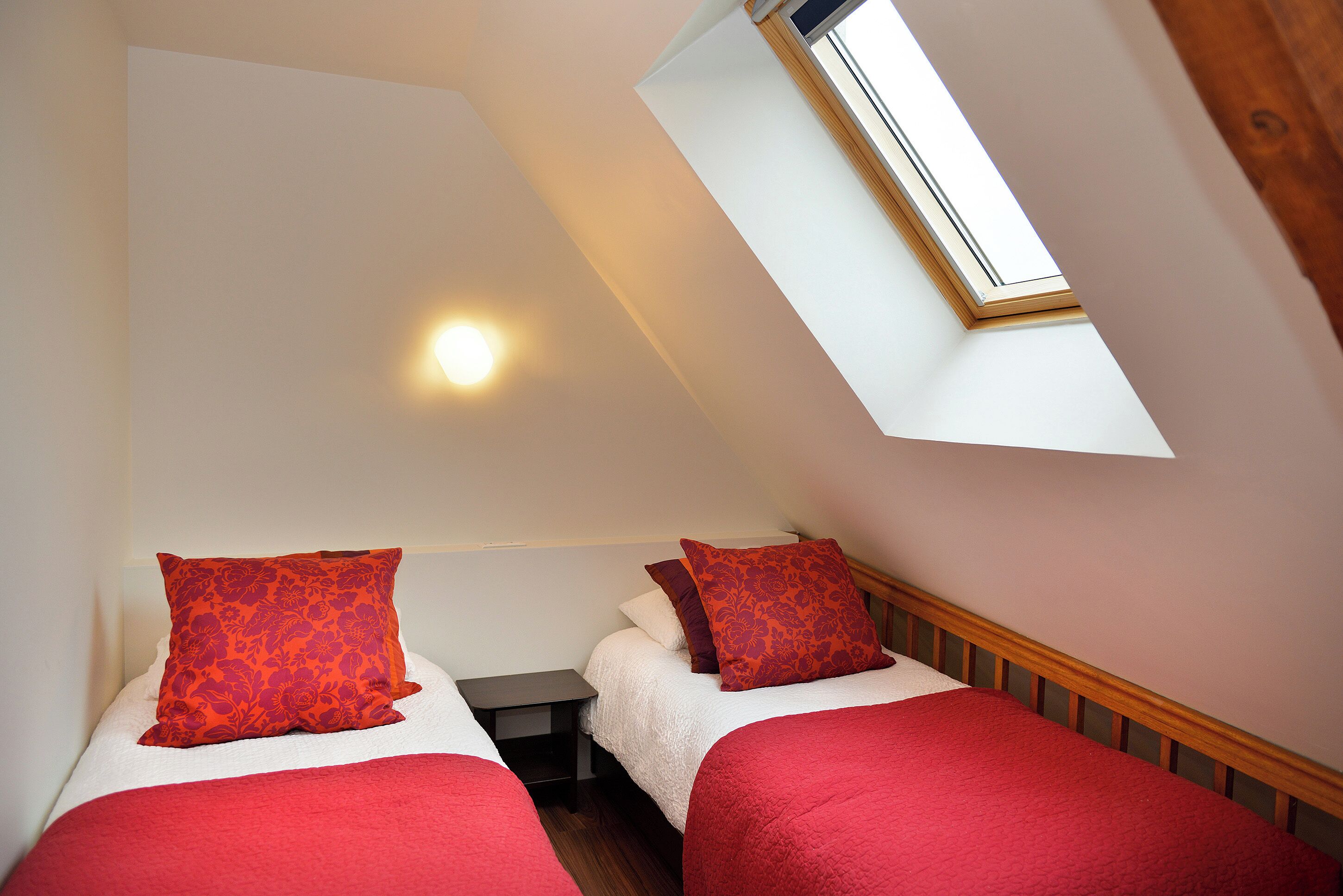 Een kamer met twee bedden, elk voorzien van rode beddengoed en kussens met bloemenprint, een klein nachtkastje en een dakraam erboven.