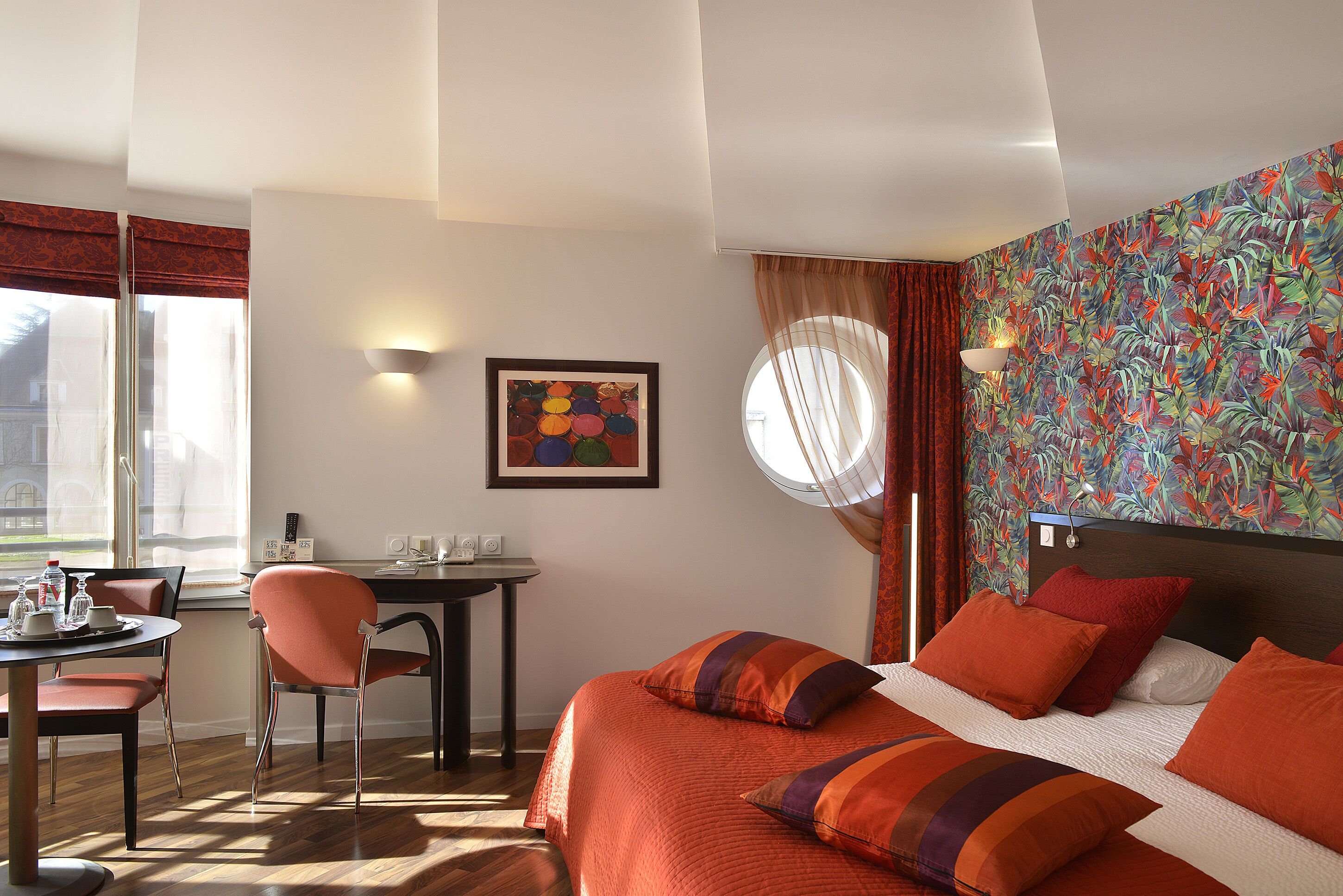 Hotelkamer met een bed, bureau en stoel. Er is een raam met gordijnen, een muur met een kleurrijke muurschildering, en een schilderij aan de wand.