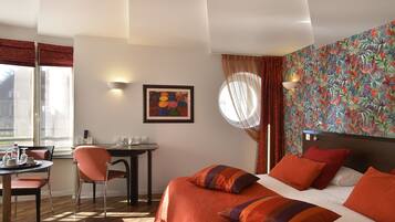 Habitación de hotel con una cama, un escritorio y una silla. Tiene una ventana con cortinas, una pared con un mural colorido y un cuadro en la pared.