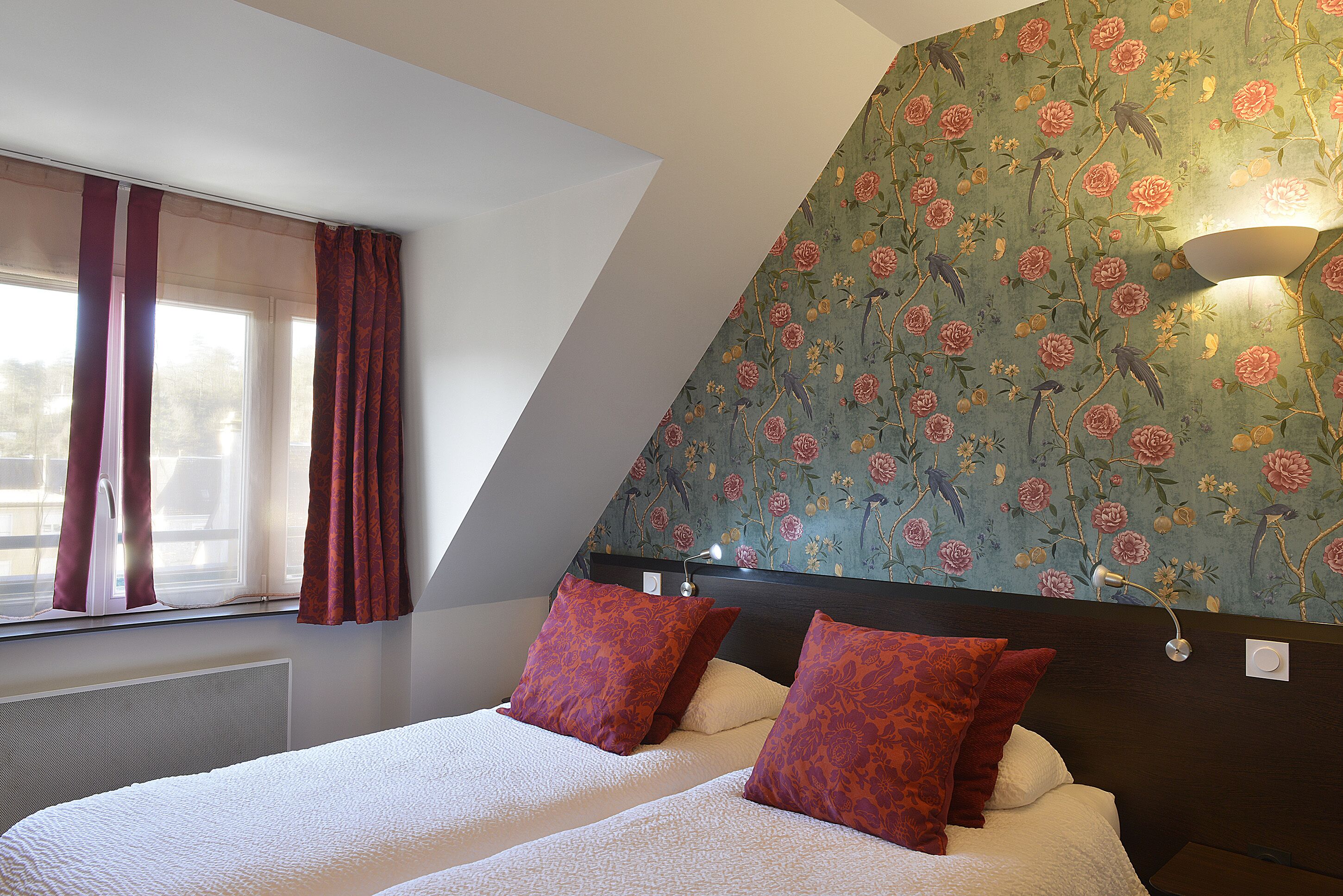 A bedroom with a large bed, two pillows, a window with curtains, and a wall with floral wallpaper.