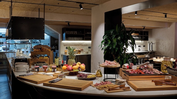 Montaje de bufé con diversos alimentos, como carnes, quesos y frutas, en una cocina bien iluminada con electrodomésticos modernos y una planta grande.