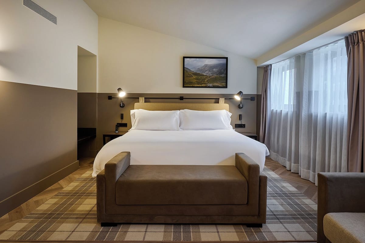 Habitación de hotel con una cama grande, cabecera con luces, vistas a montañas a través de la ventana y piso a cuadros.