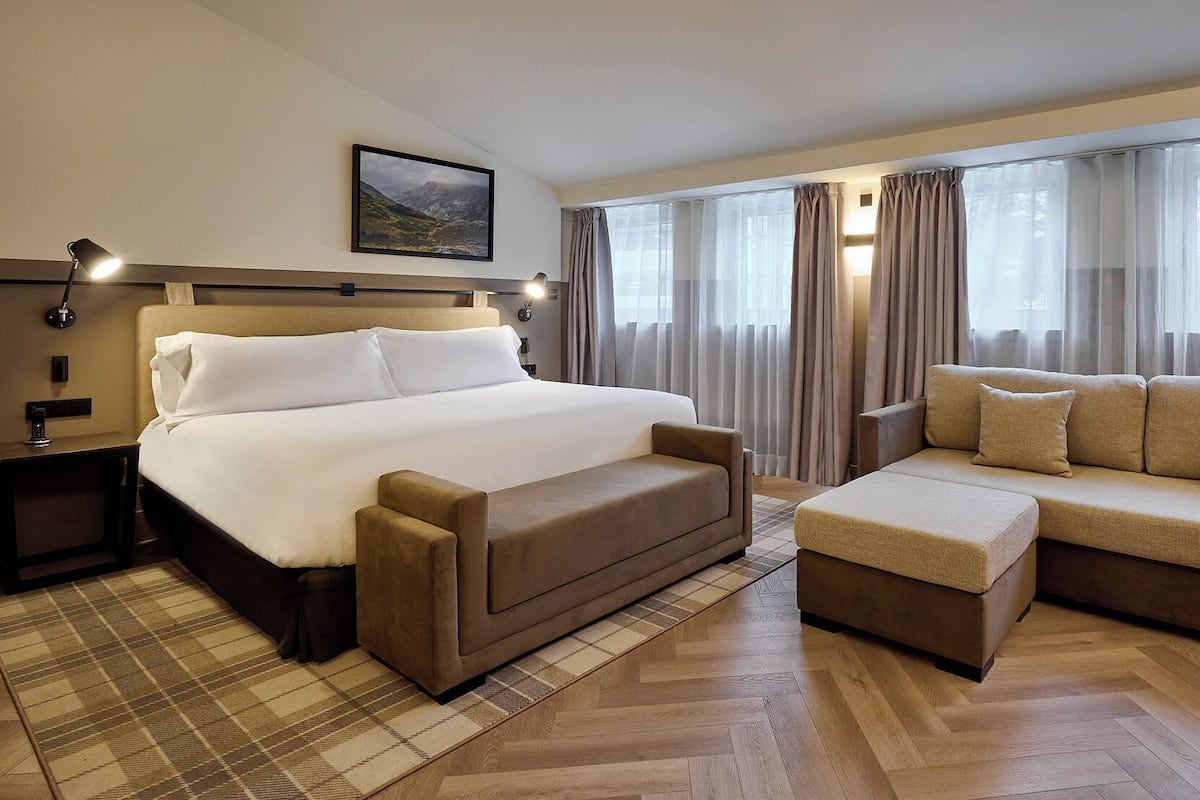Habitación de hotel con cama, sofá, mesa de centro, mesita de noche, lámpara y un cuadro en la pared.