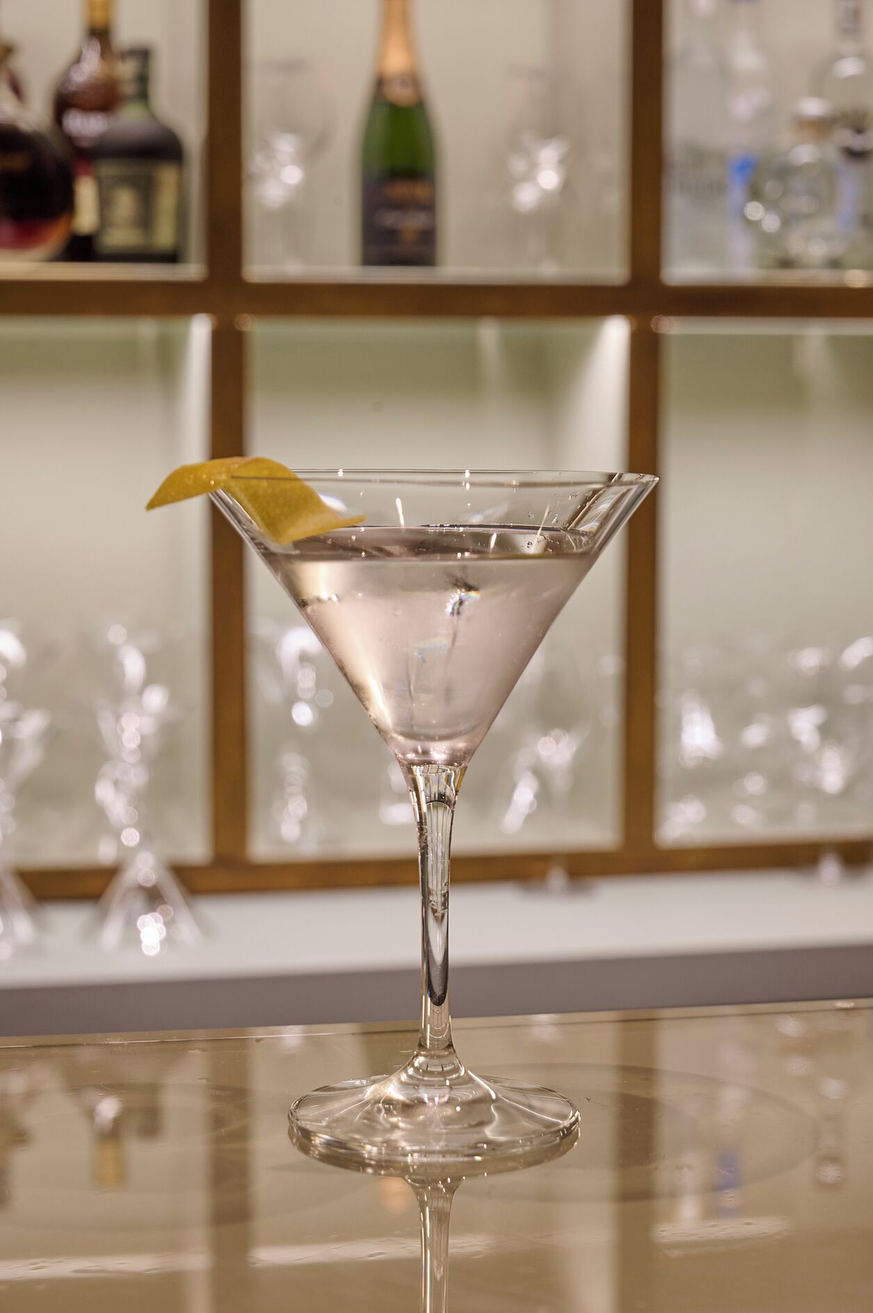 A martini glass with a lemon twist on a reflective surface.