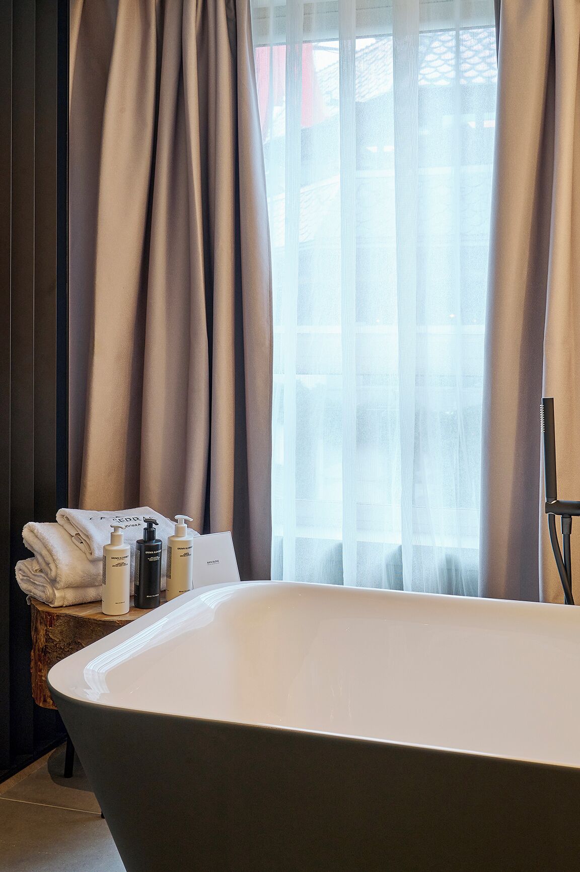 Un baño moderno con una bañera independiente, un taburete de madera con toallas y artículos de aseo, y cortinas translúcidas que dejan entrar luz natural.