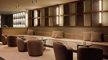 Un área de descanso moderna con sofá, sillones y pequeñas mesas, con un panel de madera con paneles de vidrio y una zona de bar con botellas.