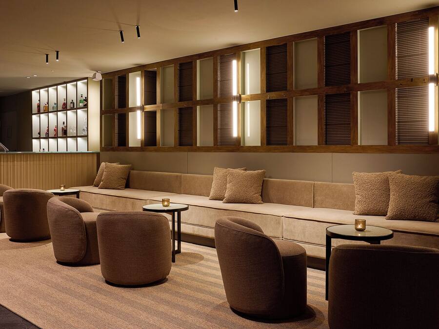 A modern lounge area with a sofa, armchairs, and small tables, featuring a wooden panel wall with glass panels and a bar area with bottles.