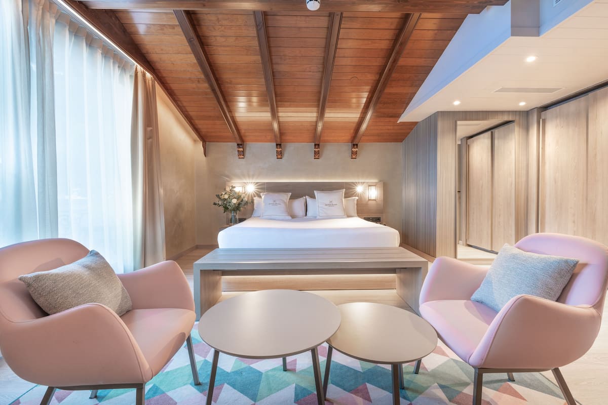 A modern hotel room with a large bed, two armchairs, a small table, and a geometric-patterned rug.