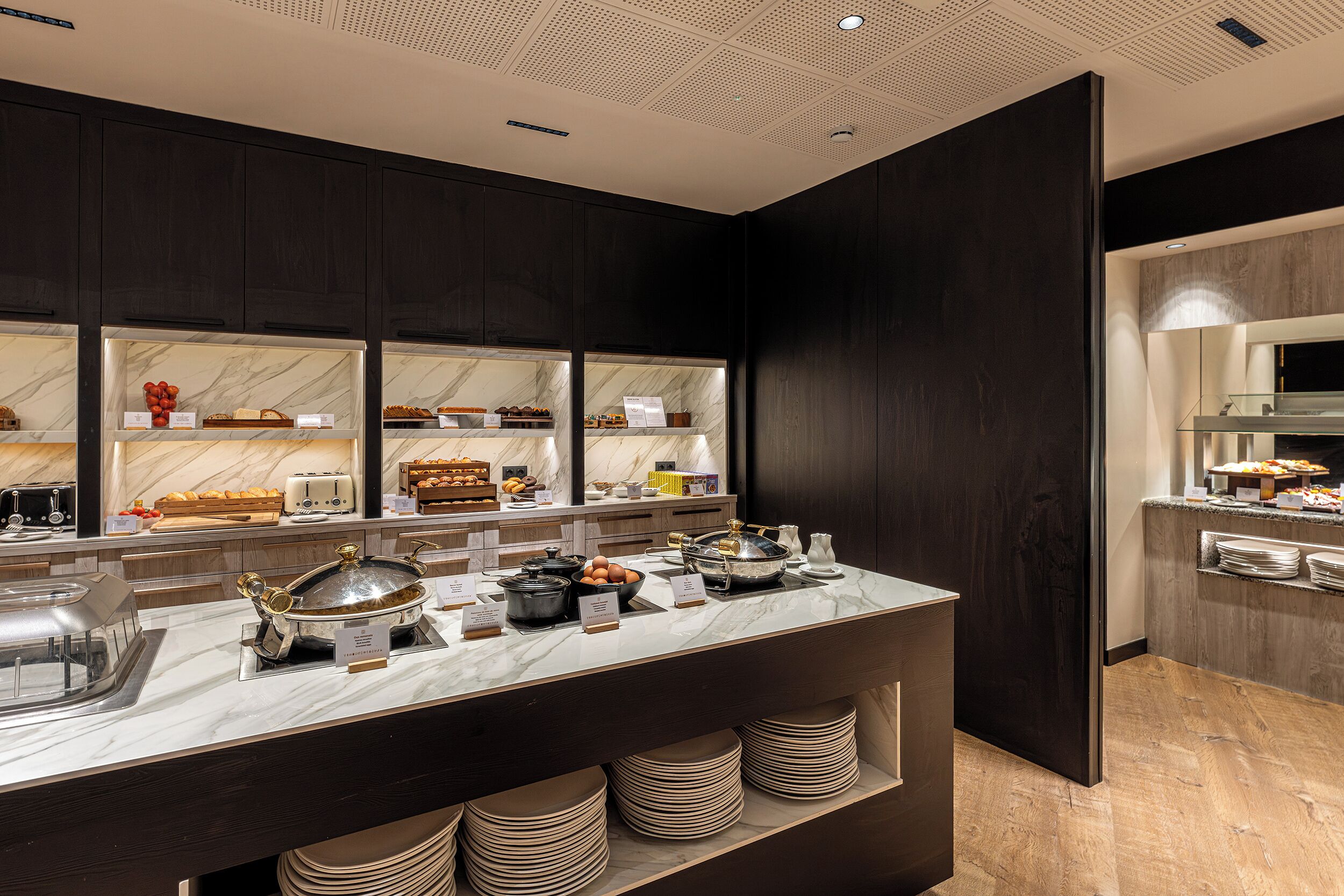 A modern kitchen with marble countertops, dark cabinetry, and a variety of food items displayed.