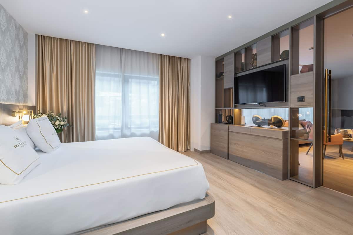 A modern hotel room with a large bed, a TV mounted on a wooden cabinet, and a seating area with a table and chairs.