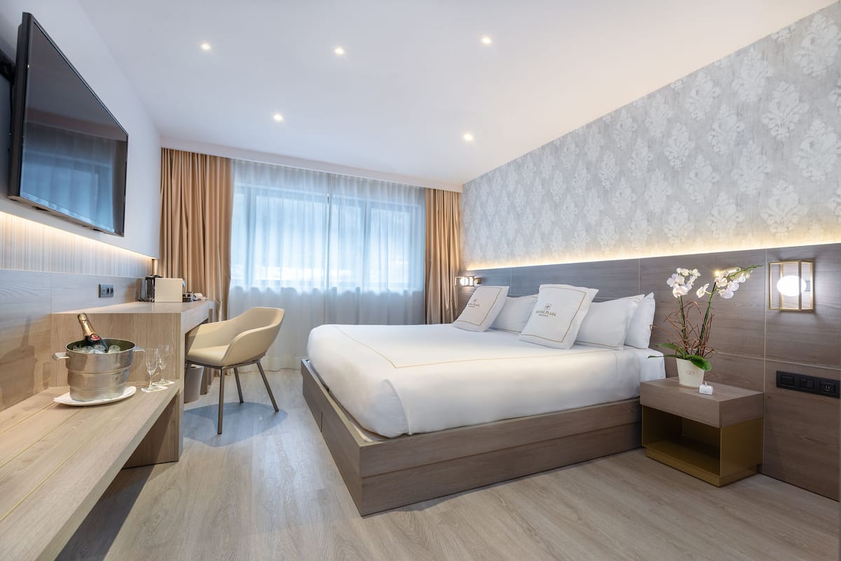 A modern hotel room with a large bed, a flat-screen TV mounted on the wall, a desk with a bottle of champagne, and a nightstand with a potted plant.