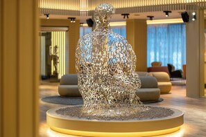 A sculpture of a human figure made of letters, placed on a circular platform in a modern lobby with seating areas and a large window.