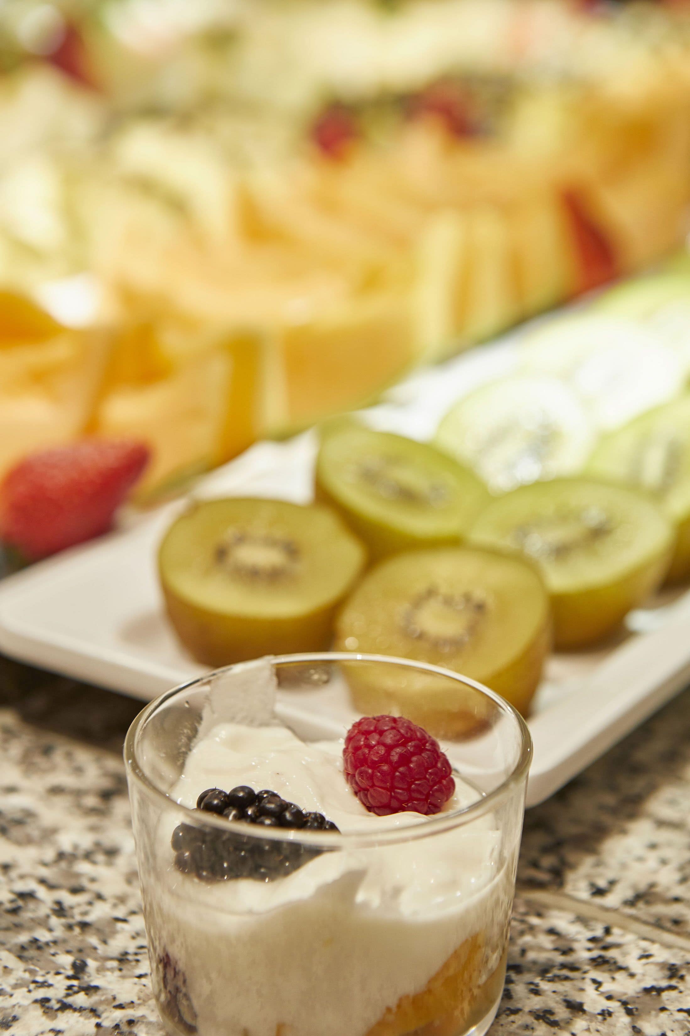 Un vaso de postre con crema batida y frutos rojos, un plato de frutas en rodajas y un fondo desenfocado con más frutas.