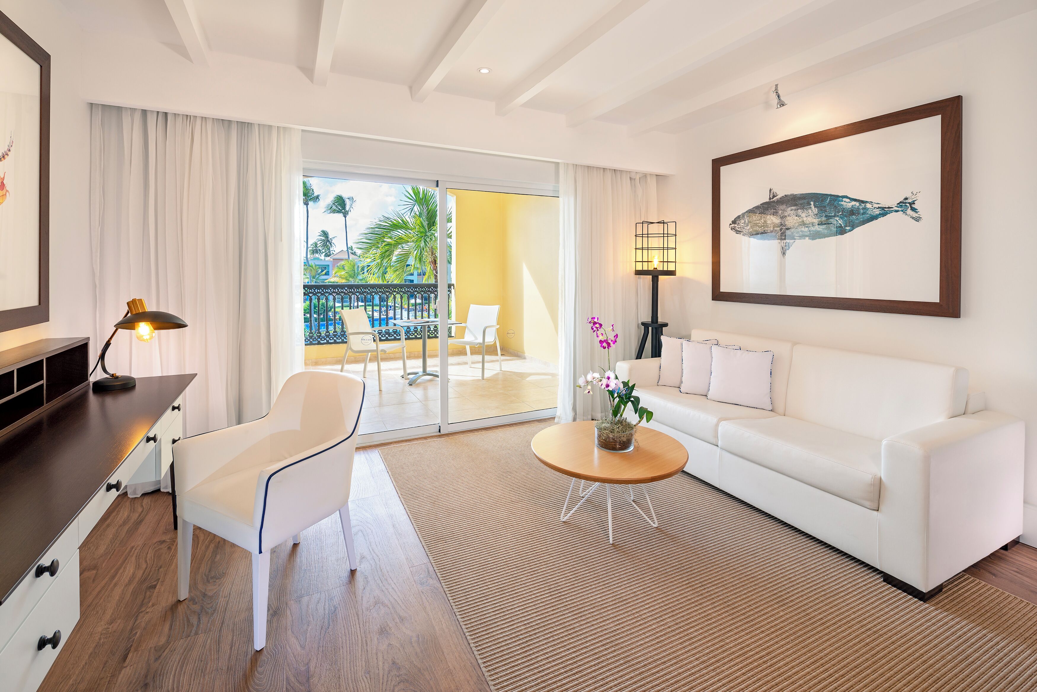 A modern hotel room with a white sofa, a white armchair, a wooden coffee table with a flower vase, a desk with a lamp, and a large window with a view of a balcony and palm trees.
