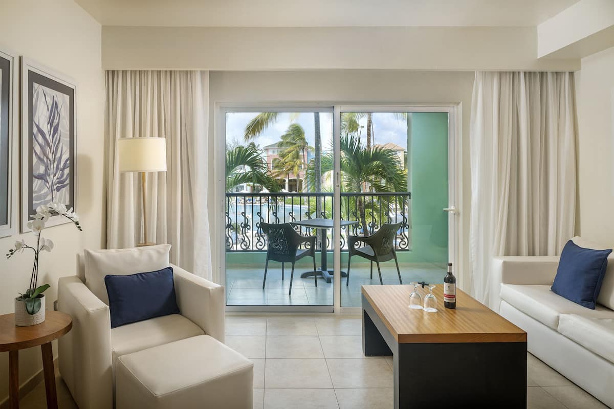 A hotel room with a balcony, a sofa, a chair, a coffee table, and a lamp.