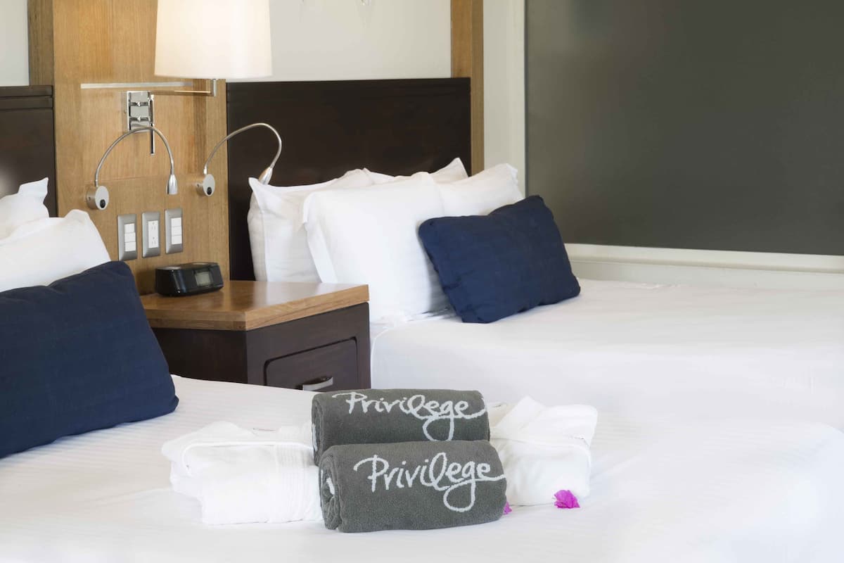 A hotel room with a neatly made bed, two towels with 'Privilege' branding, a bedside table with a lamp, and a nightstand with a phone and a small appliance.