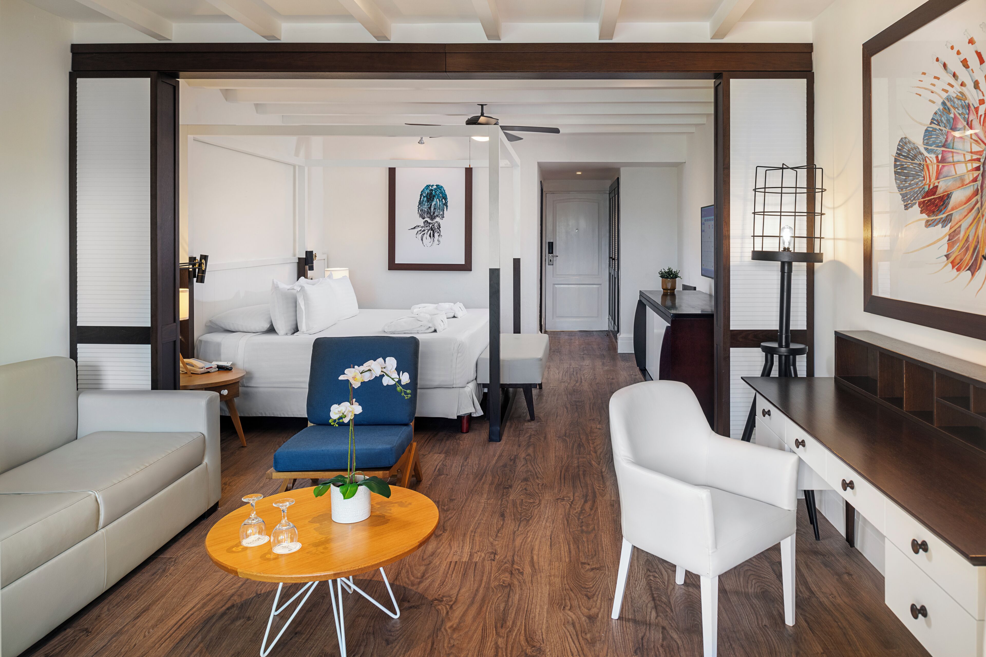 Ein modernes Hotelzimmer mit einem Bett, einem blauen Sessel, einem weißen Sofa, einem Holztisch und einem Schreibtisch mit Schubladen.
