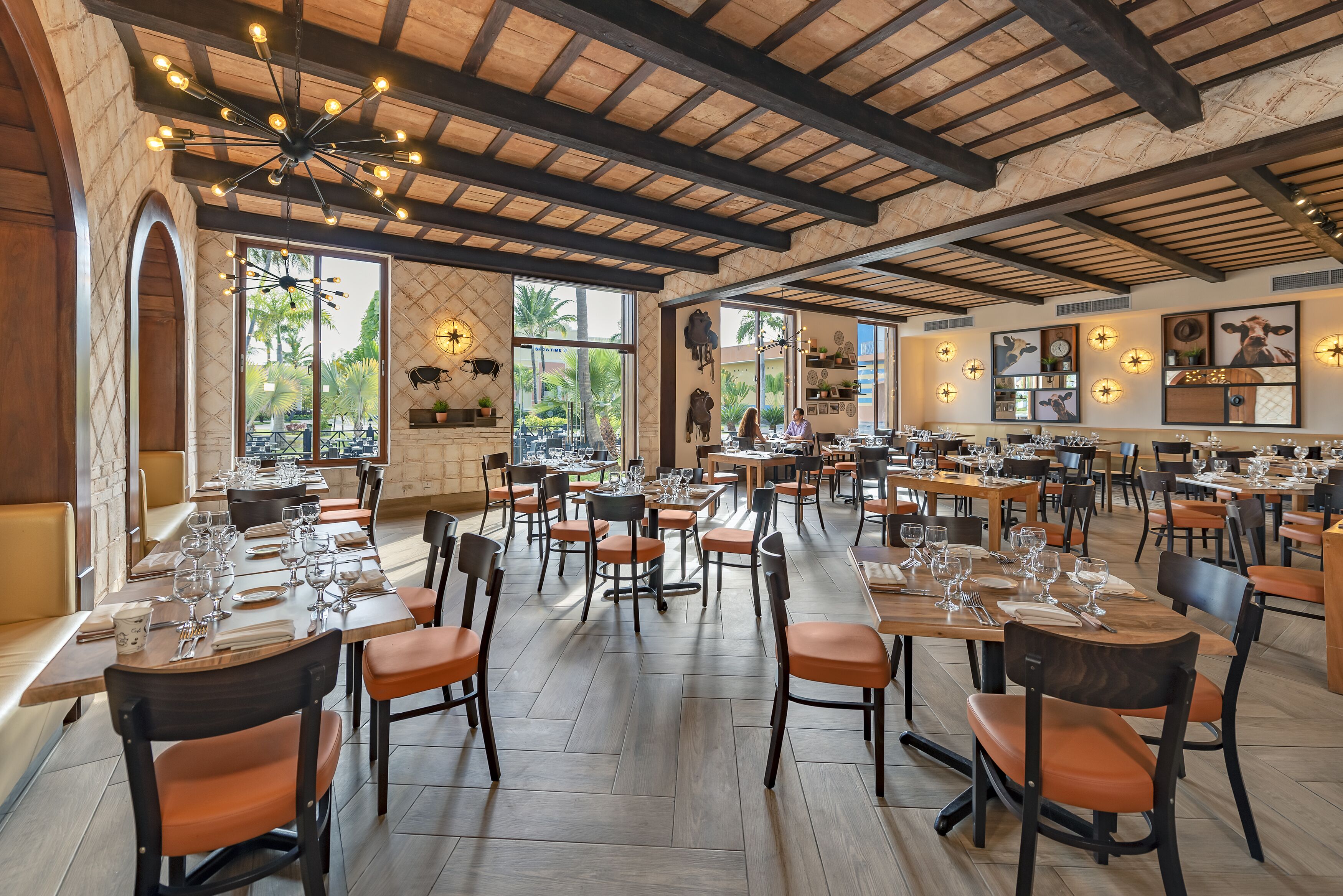 A spacious restaurant with high ceilings, exposed beams, and large windows offering views of palm trees. Tables are neatly set with wine glasses and plates.