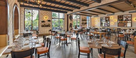 A spacious restaurant with high ceilings, exposed beams, and large windows offering views of palm trees. Tables are neatly set with wine glasses and plates.